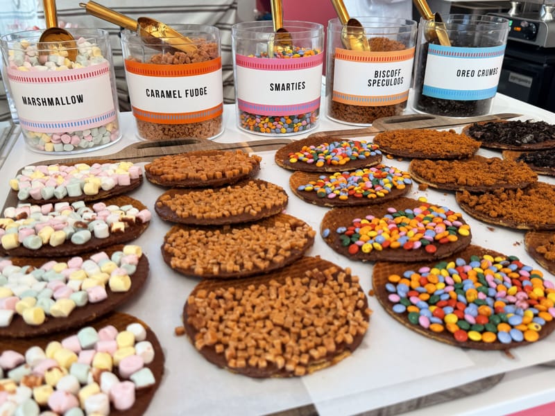 Five labelled jars of toppings on the counter, marshmallow, caramel fudge, smarties, Biscoff speculoos and Oreo crumbs, with gold scoops, above a tray of finished chocolate stroopwafels